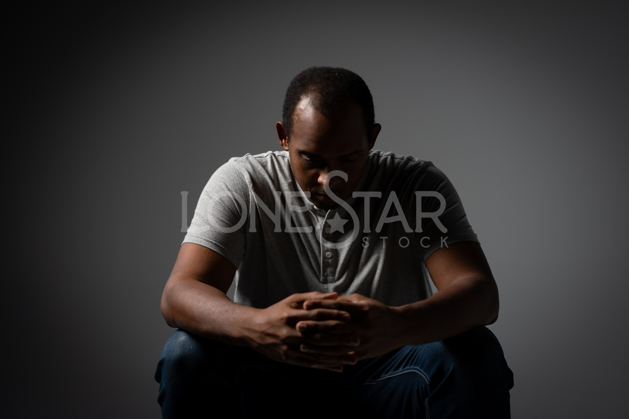 Preview of Depressed Black Man in a gray shirt Sitting and Looking Down and holding his hands together